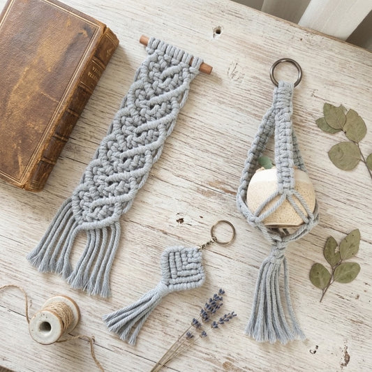 A styled flat-lay of a light gray macramé keychain holder with a wooden bead and metal key ring, shown alongside a matching knotted tassel bookmark on a rustic white wooden tabletop with a vintage book, thread spool, dried lavender, and green leaves.