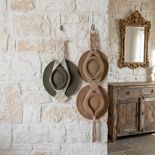 Two wide-brimmed hats hanging from macrame wall hangers on a rustic white stone wall, with a wooden cabinet and ornate mirror in the background.