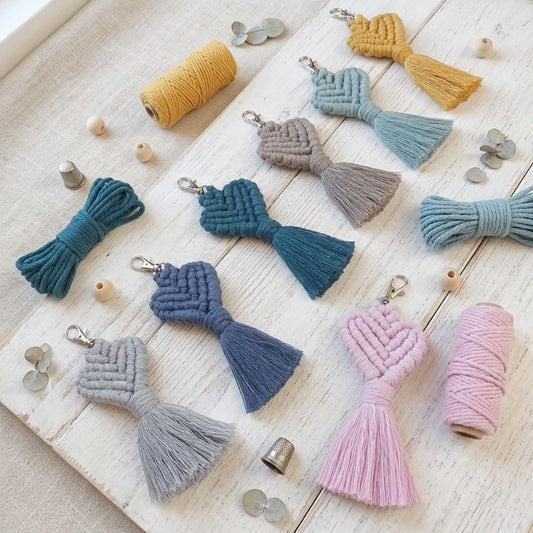 A flat-lay display of handmade macrame tassel keychains in various colours (blue, pink, grey, teal, mustard, and light blue), shown with their matching thread spools on a whitewashed wooden background.