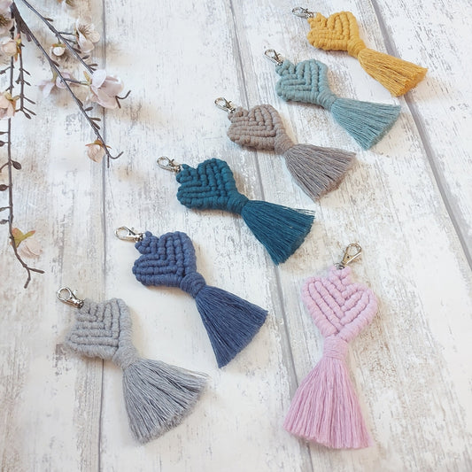 A variety of handmade macrame keyrings in different colours displayed on a wooden surface, each shaped like a heart with a loop at the top for attaching to keys.