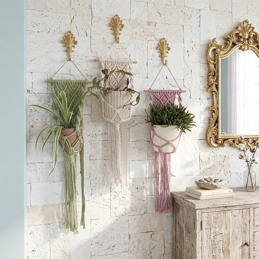 Three bohemian macrame wall hangings in green, cream, and dusty pink, each holding a potted plant, displayed on a white brick wall next to a gold mirror and wooden cabinet.
