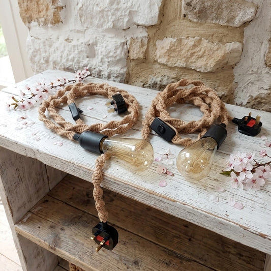 Two sets of rustic woven macrame cord string lights with black switches and UK plugs, displayed on a distressed white wooden table with cherry blossoms and a stone wall background.