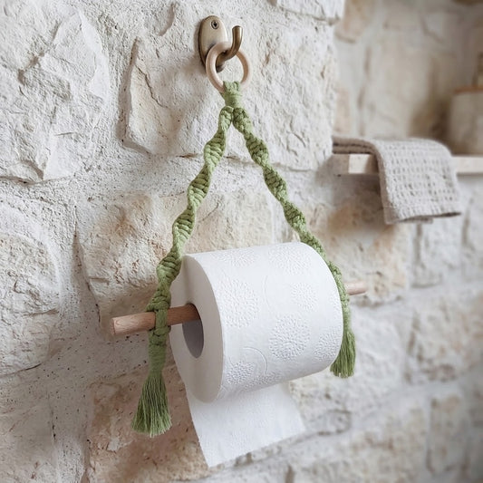 A green braided rope toilet paper holder with tassels and a wooden roll holder, holding a white toilet paper roll on a rustic white stone wall.