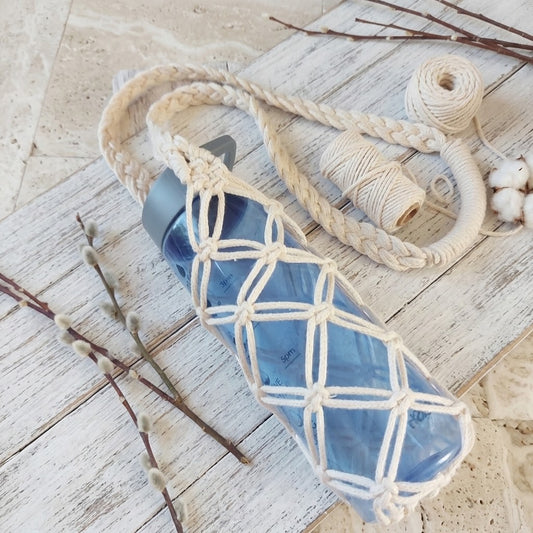 A translucent blue water bottle wrapped in a cream macrame carrier with a matching braided shoulder strap, styled on a whitewashed wooden surface with twigs, cotton bolls, and spools of thread.