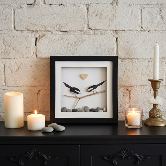 A framed shadow box artwork featuring two magpies perched on a branch with a small gold heart, displayed on a dark wooden cabinet against a white brick wall, styled with lit candles.