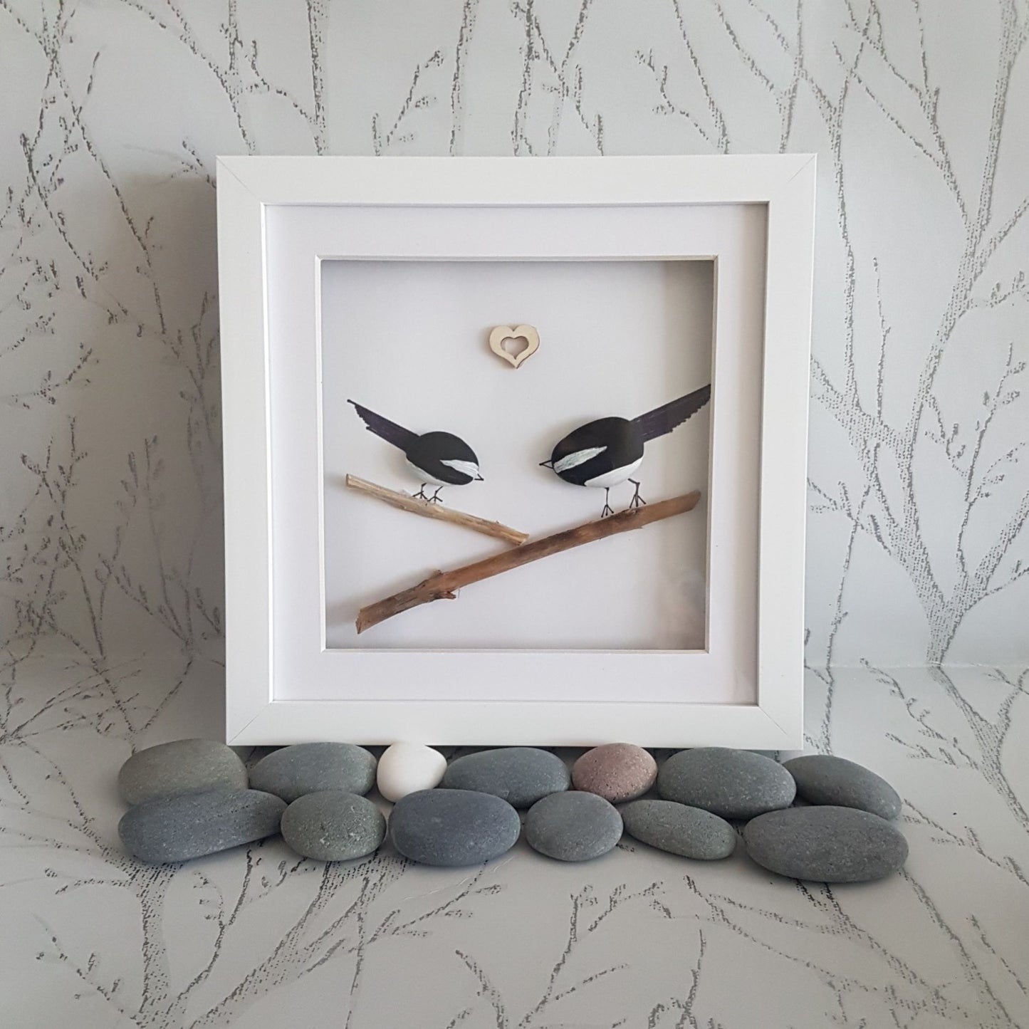 A pebble art picture frame containing a picture of two magpies, with a heart-shaped cut-out in the center, displayed on a bed of pebbles.