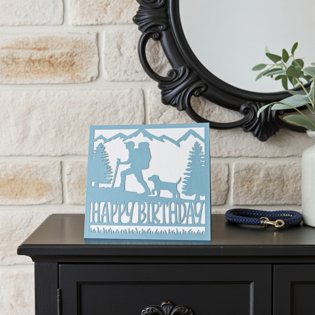 A square light blue and white 'Happy Birthday' card featuring a silhouette of a hiking backpacker and dog, displayed on a dark wooden cabinet against a light stone wall with a black mirror and a plant.