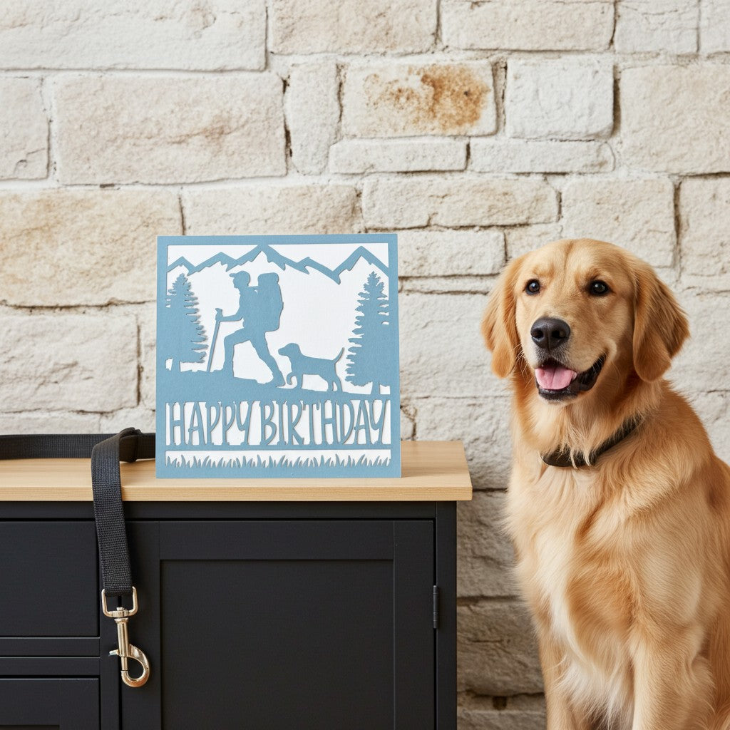 A square light blue and white 'Happy Birthday' card with a hiking dog silhouette, displayed on a wooden cabinet next to a golden retriever dog against a light brick wall.