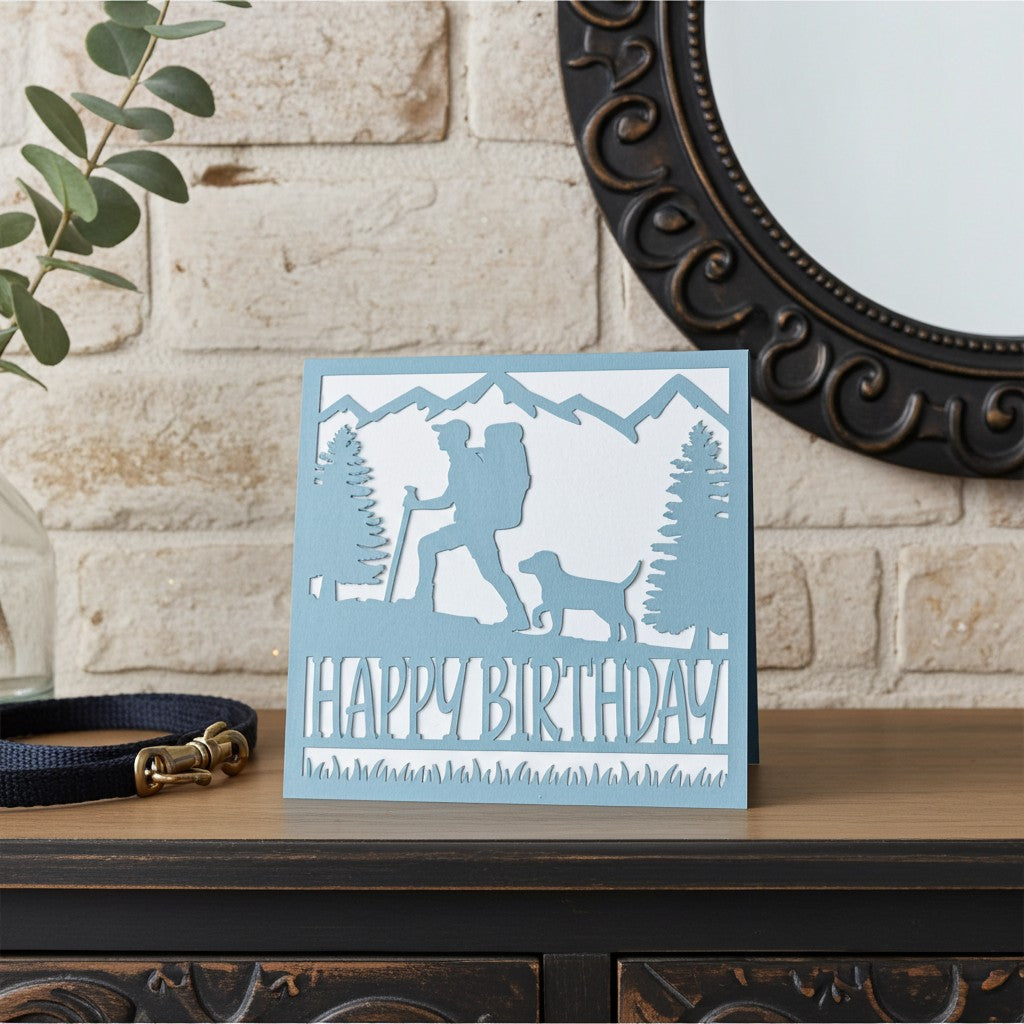A square birthday card with a light blue and white laser-cut outdoor scene featuring a hiker, dog, trees, and mountains, with the words “HAPPY BIRTHDAY” across the bottom. The card is styled on a wooden surface in front of a light brick wall, with a dark mirror frame and a navy pet collar nearby.