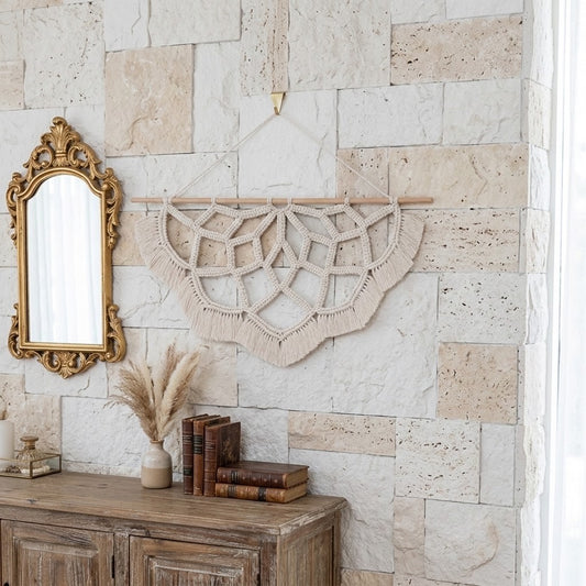 A bohemian-style macrame wall hanging in a warm beige color, suspended from a wooden dowel on a stone wall above a rustic wooden console table with books and a mirror.