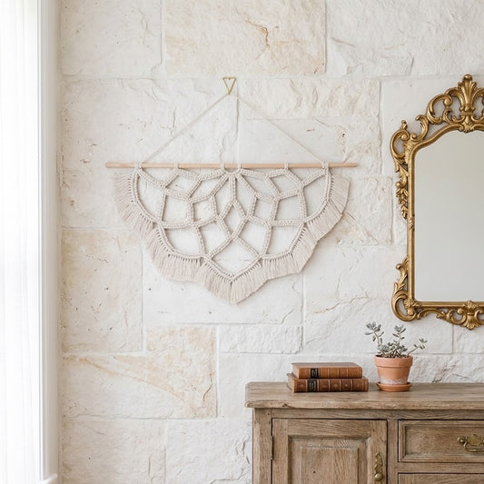A cream macrame wall hanging with intricate geometric woven patterns and fringe, suspended from a light wooden dowel on a textured beige stone wall above a rustic wooden console table with books and a potted plant.