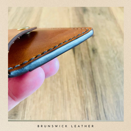 Brown leather wallet with silver edge held by a hand on a wooden surface, branded 'Brunswick Leather'.