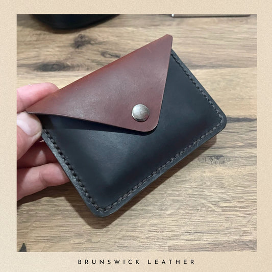 Hand holding a leather wallet with a brown flap on a wooden surface, branded 'Brunswick Leather'.