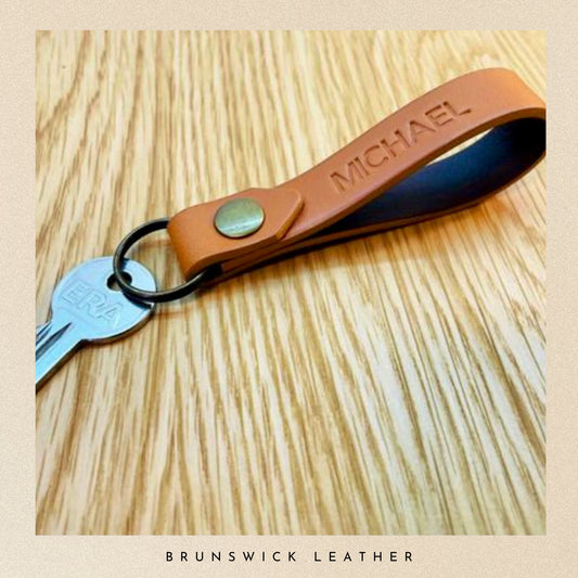 Brown leather keychain with 'MICHAEL' engraving on a wooden surface, branded 'Brunswick Leather'.