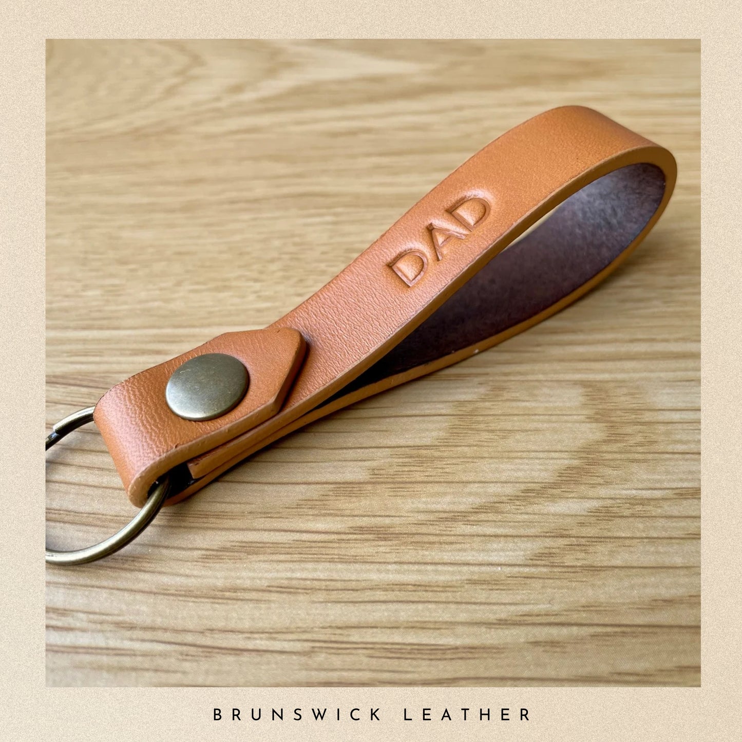 Brown leather keychain with 'DAD' engraving on a wooden surface, branded 'Brunswick Leather'.