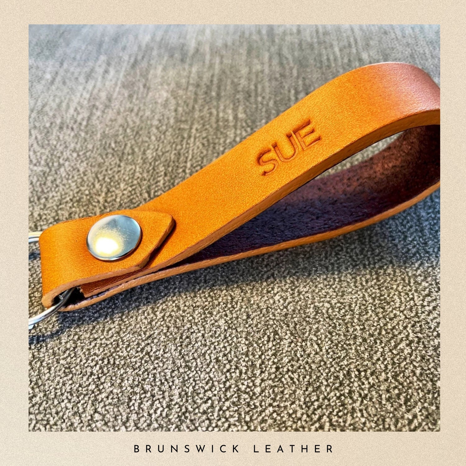 Brown leather key fob with 'SUE' engraving on a textured surface, branded 'Brunswick Leather'.