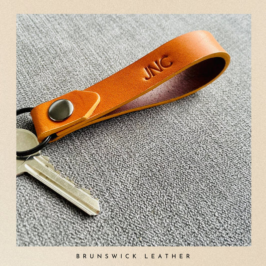 Brown leather keychain with 'JNC' engraving on a textured surface, branded 'Brunswick Leather'.