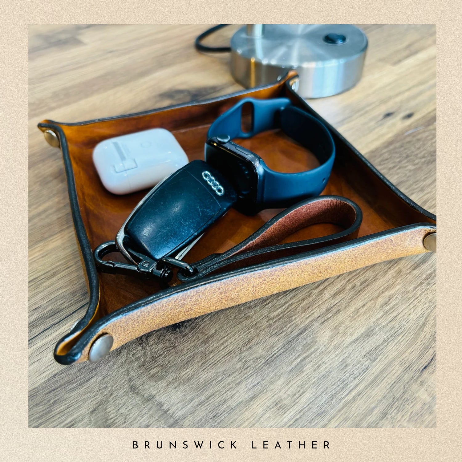 Brown leather tray with electronic devices on a wooden surface, branded 'Brunswick Leather'.