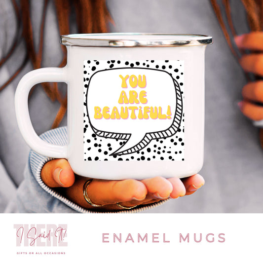 A white ceramic enamel mug with black and yellow text pattern, held in a person's hand. The text on the mug reads 'YOU ARE BEAUTIFUL!' in a stylized font, and the mug is presented with a 'THERE I SAID IT' branding.