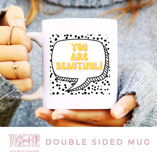 A person holding a white double-sided mug with a black and yellow speech bubble graphic that says 'You are beautiful!' and small dots pattern around it.