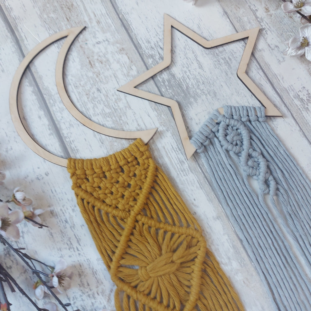 Two macrame wall hangings, one shaped like a crescent moon and the other resembling a star, in beige and brown colours, displayed on a white wooden background with a floral arrangement and a background of a white fence and flowers.