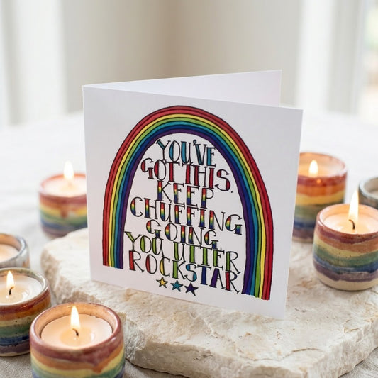 A white greeting card with a coloUrful rainbow and the text 'YOU'VE GOT THIS KEEP CHUFFING GOING YOU UTTER ROCKSTAR' stands on a stone surface surrounded by lit rainbow candles.