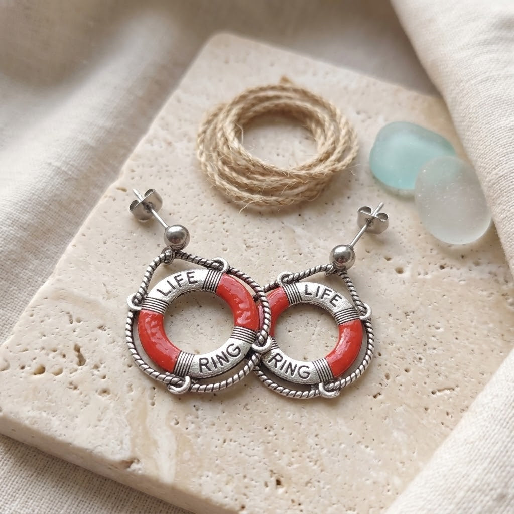 A pair of silver-toned hoop earrings with red life preserver ring details and the words 'LIFE RING' engraved on each hoop, styled on a beige stone slab with twine and sea glass.