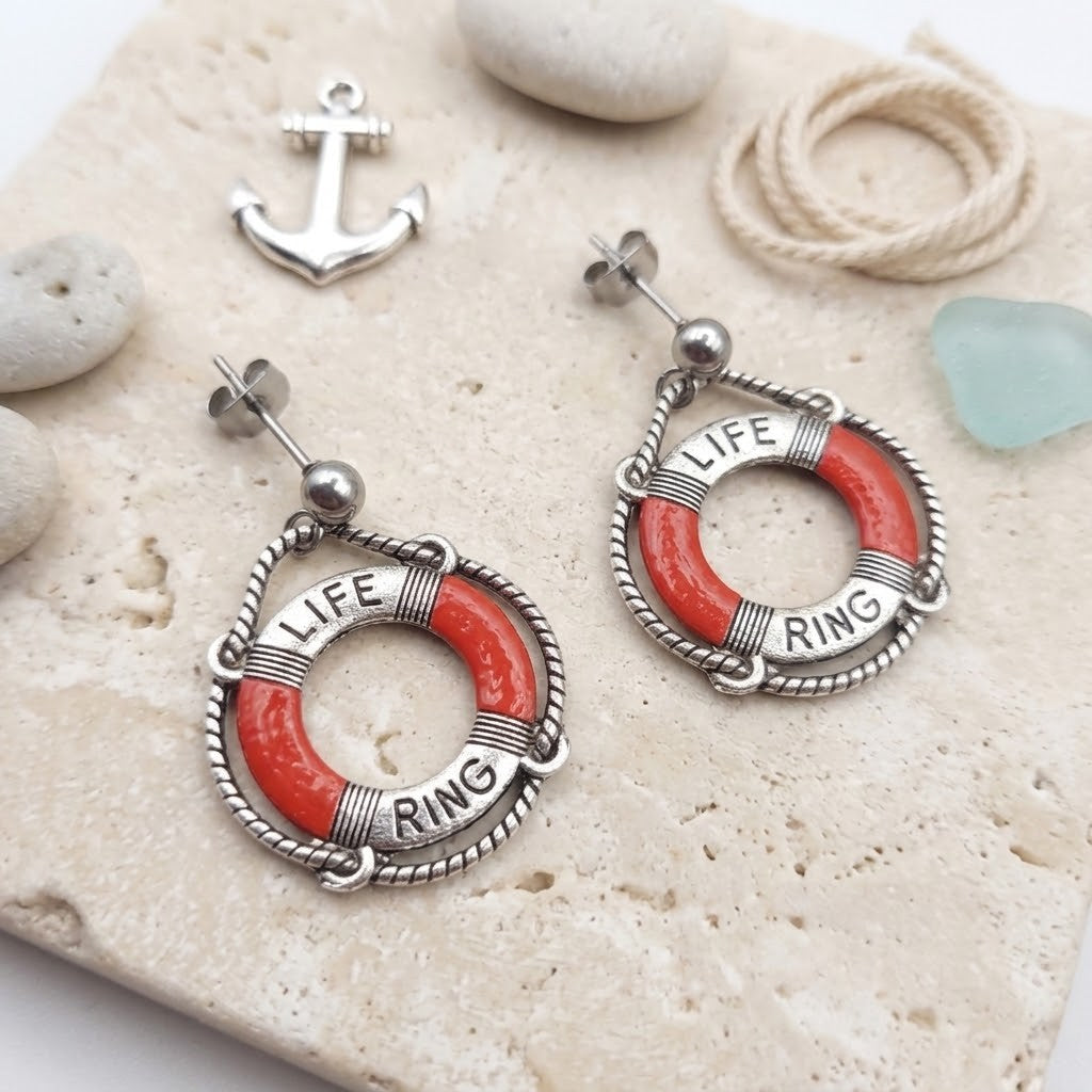 A pair of silver-toned life ring dangle earrings with coral-red enamel sections and the words 'LIFE RING' engraved, displayed on a light stone surface with small nautical props including an anchor charm, rope, and sea glass.