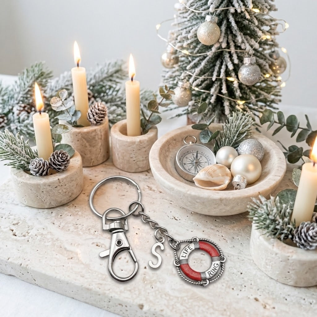 A silver-toned metal keychain with a lobster clasp, a letter S charm, and a red-and-silver life preserver charm engraved with 'LIFE RING', displayed on a stone surface with candles, pinecones, eucalyptus, and a decorated Christmas tree in the background.