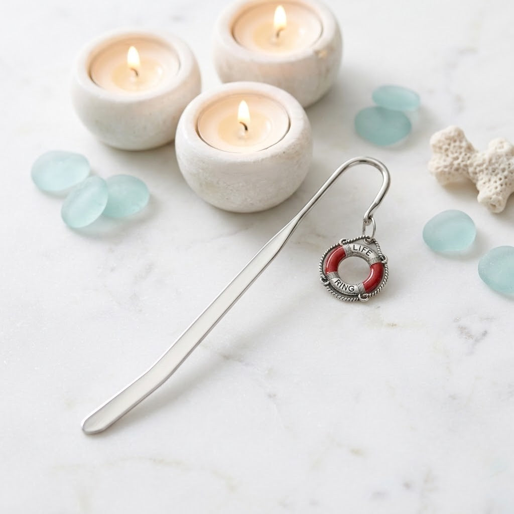 A silver metal bookmark shaped like a life preserver ring with red and white enamel detailing, displayed on a white marble surface with lit candles, sea glass, and coral as props.