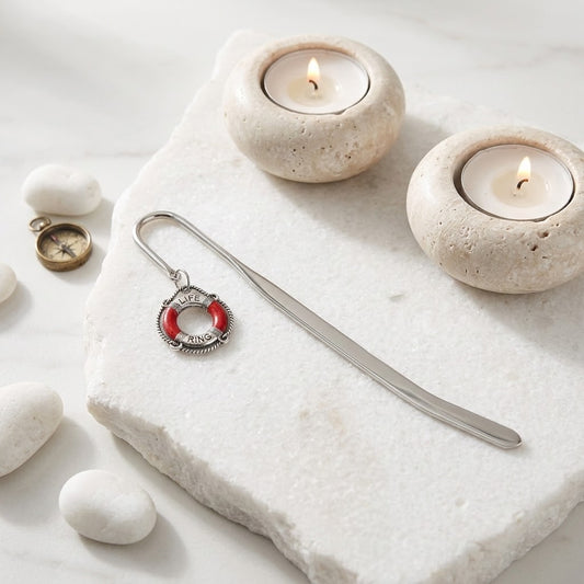 A silver metal bookmark with a red life preserver charm displayed on a white stone slab alongside two lit tealight candles, white stones, and a small brass compass.