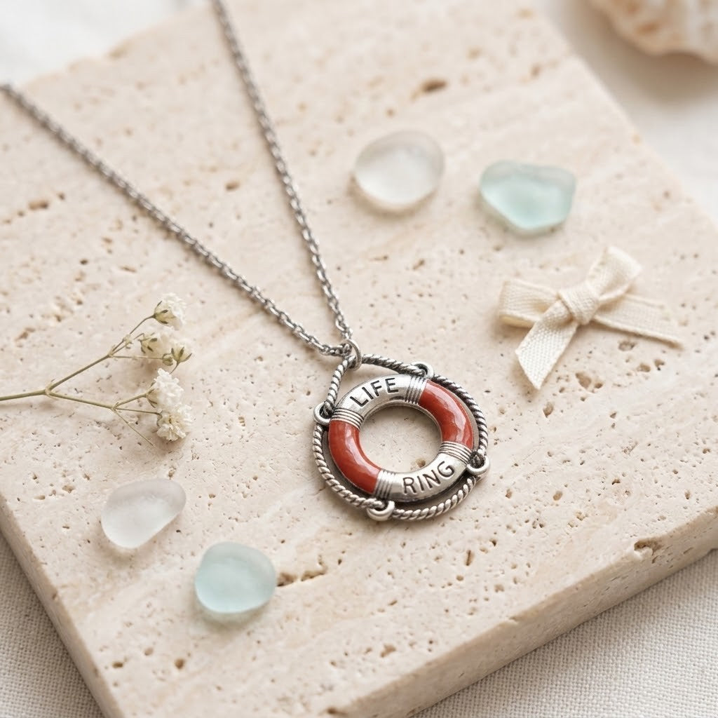 A silver chain necklace with a life preserver pendant featuring red and white styled on a textured stone tile with small sea glass and dried flowers.