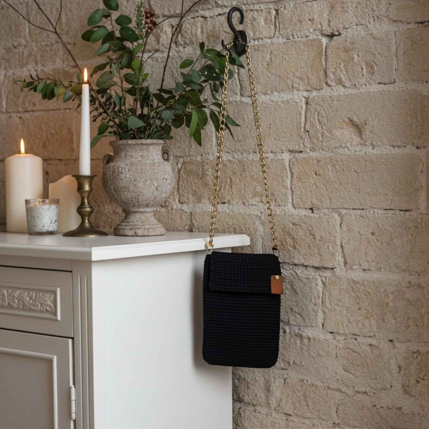 Navy handbag with a gold chain strap hanging on a hook against a stone wall with candles and a vase.