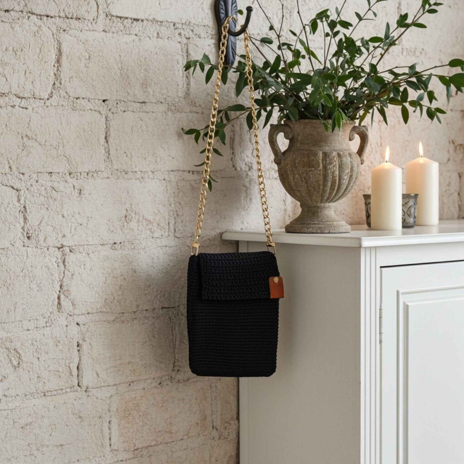 Navy woven handbag with a gold chain strap hanging on a brick wall next to a white cabinet with candles and a vase.