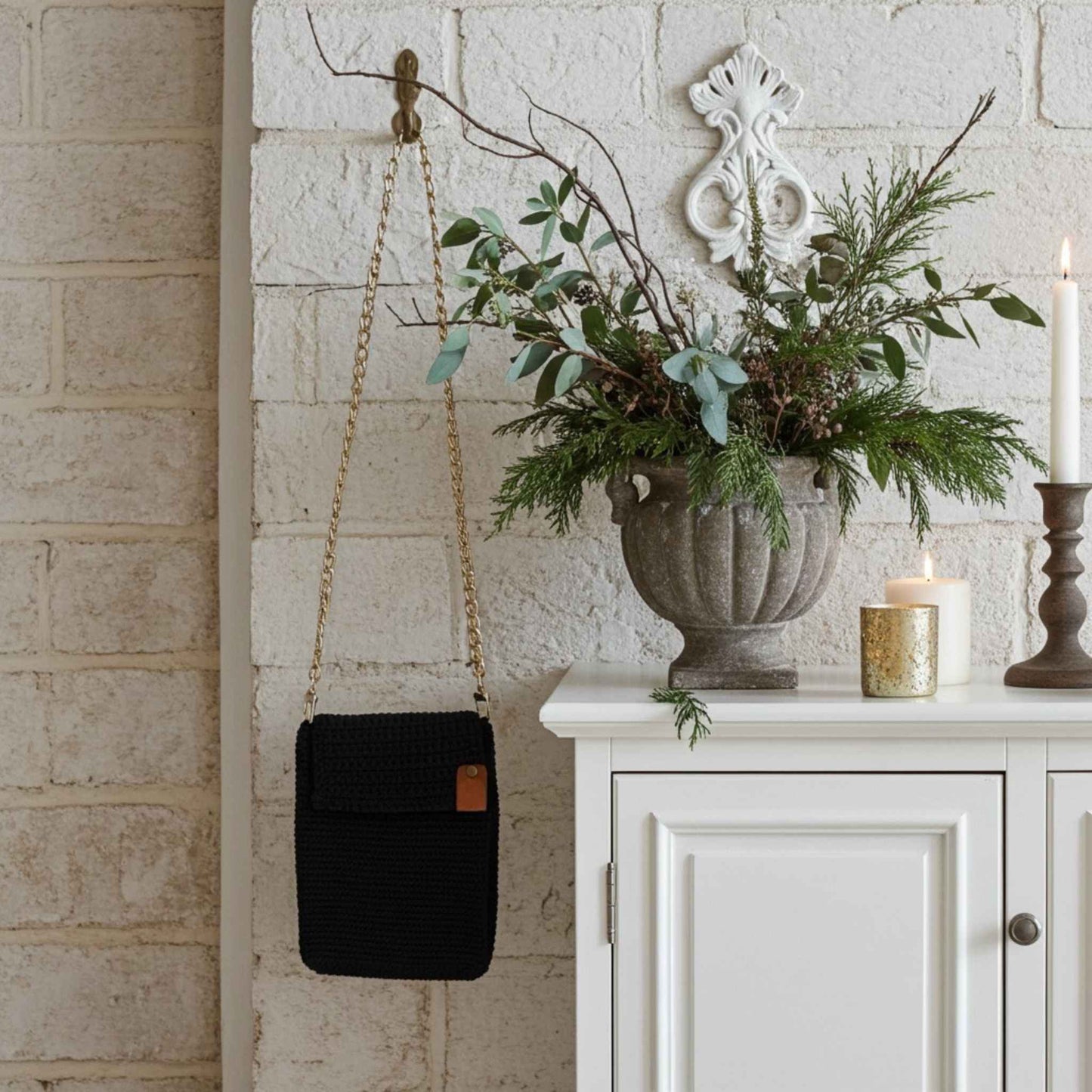 Decorative setting with a navy phone bag, potted plant, and candles against a white brick wall.