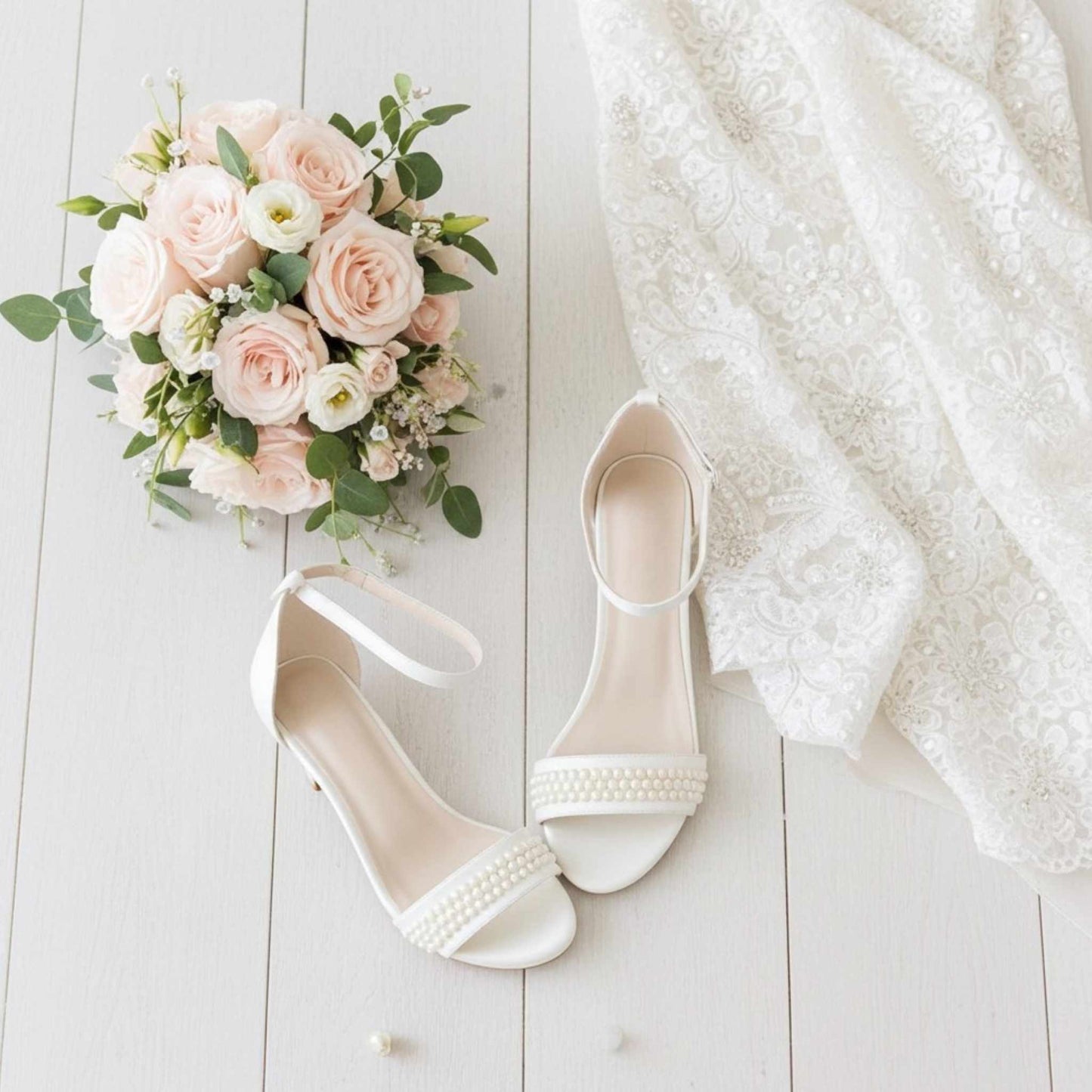 Ivory wedding shoes with pearls, a bouquet of pink and white roses, and a white lace garment on a light wooden surface.