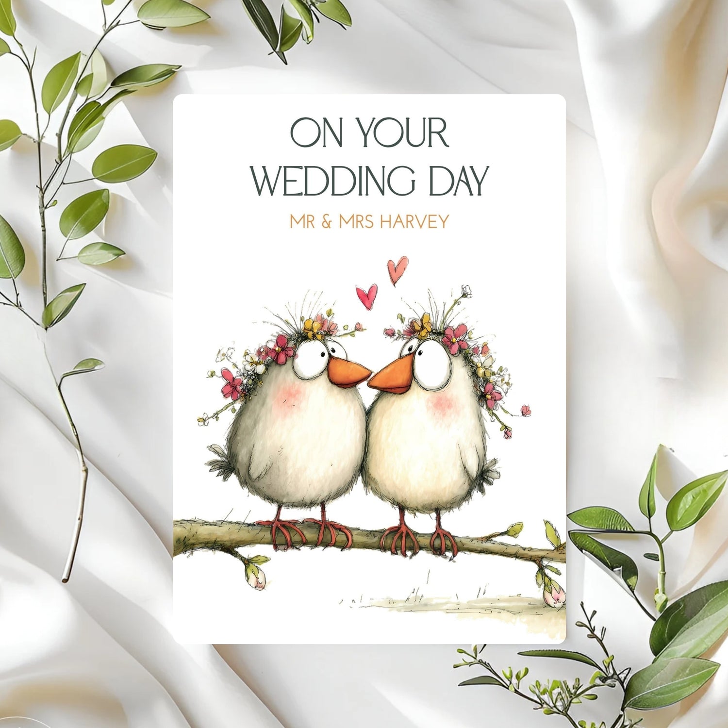 Wedding card with two birds on a branch, surrounded by green leaves on a white fabric background.