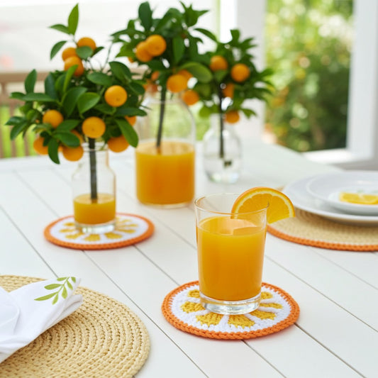 A styled dining table scene with three clear glass bottles and a glass of orange juice arranged with small orange-and-white coasters, woven placemats, plates, and leafy orange branches in the background.