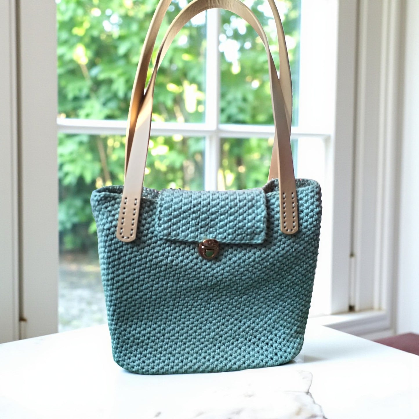 crochet shoulder bag with beige handles on a white surface with a blurred green outdoor background