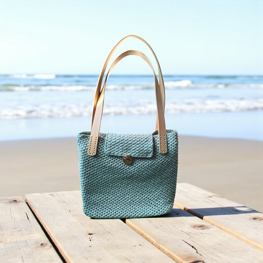 crochet shoulder bag standing on a rustic table with the sea and sandy beach in the background