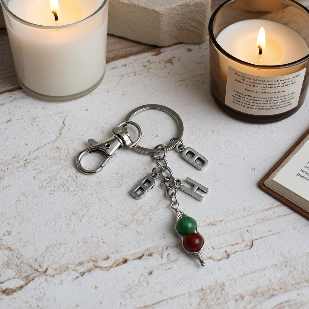A silver-toned metal keychain with a lobster clasp, three small rectangular charms, and a dangling beaded charm in green and red, styled on a light textured tabletop with lit candles in the background.