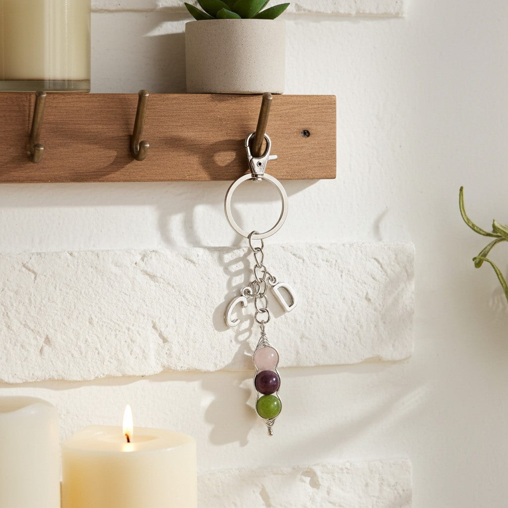A silver keychain with a key ring, small charms, and three stacked gemstone beads in pink, purple, and green, hanging from a wooden coat rack against a white brick wall with candles nearby.