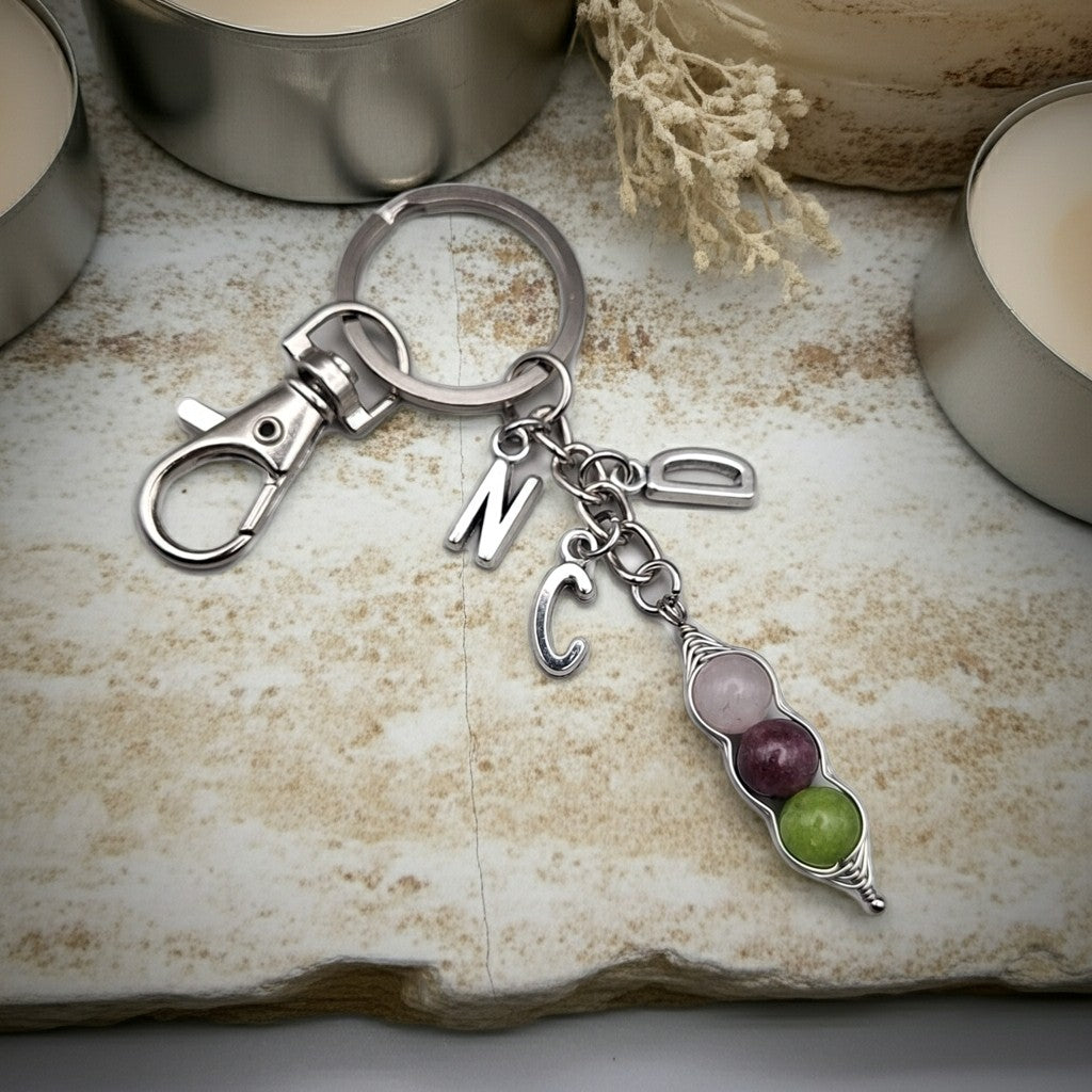 A silver-toned keychain with a lobster clasp, split ring, and dangling charms including a letter 'N', a letter 'C', a small heart, and a pebble charm with pink, purple, and green stones, displayed on a textured stone surface with candles in the background.