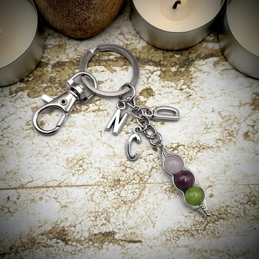A silver-toned keychain with a lobster clasp, split ring, and dangling charms including letters and a small beaded charm in pale pink, purple, and green, displayed on a textured stone surface with blurred tealights in the background.