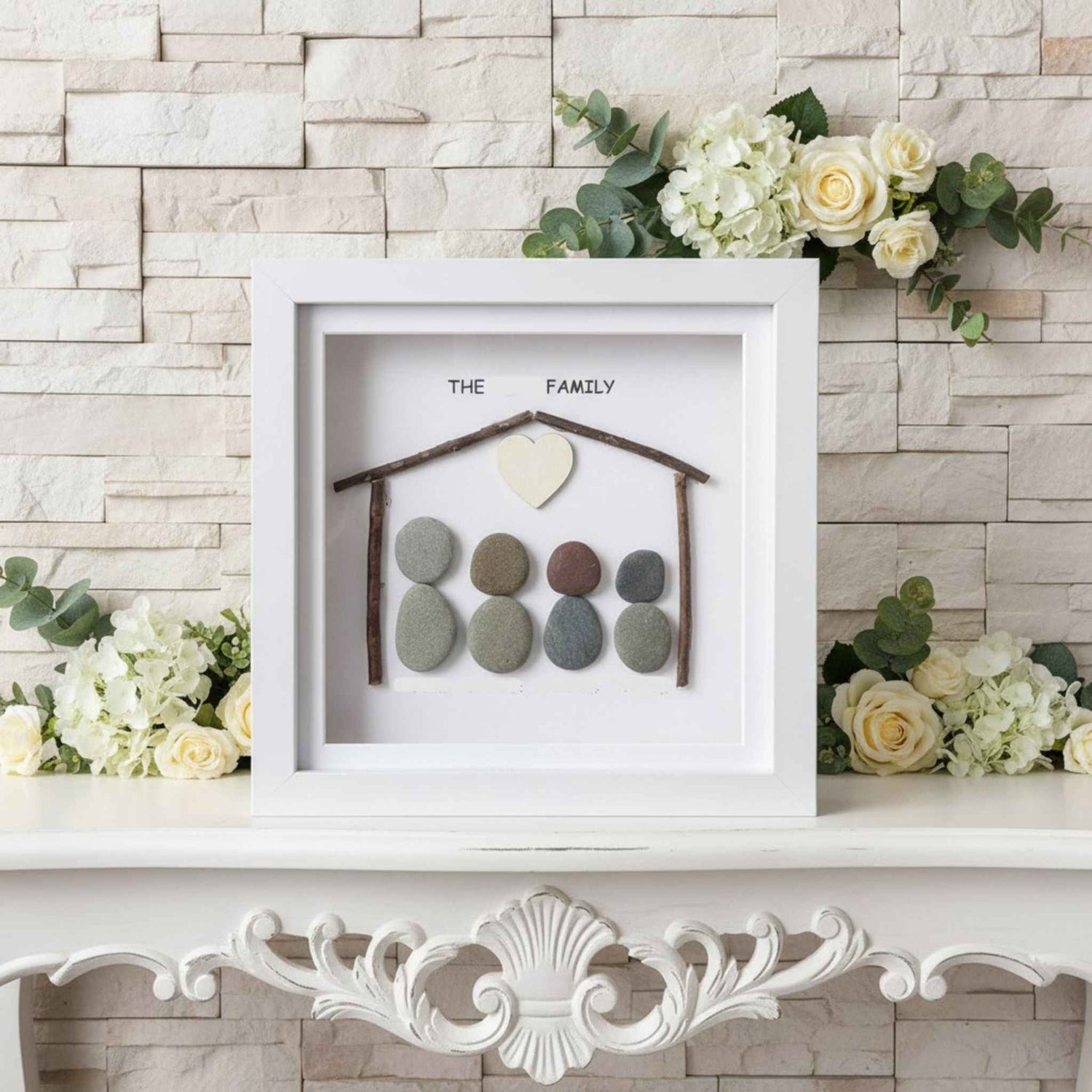 Decorative wall art with pebble house and 'The Family' text on a white frame, surrounded by flowers.