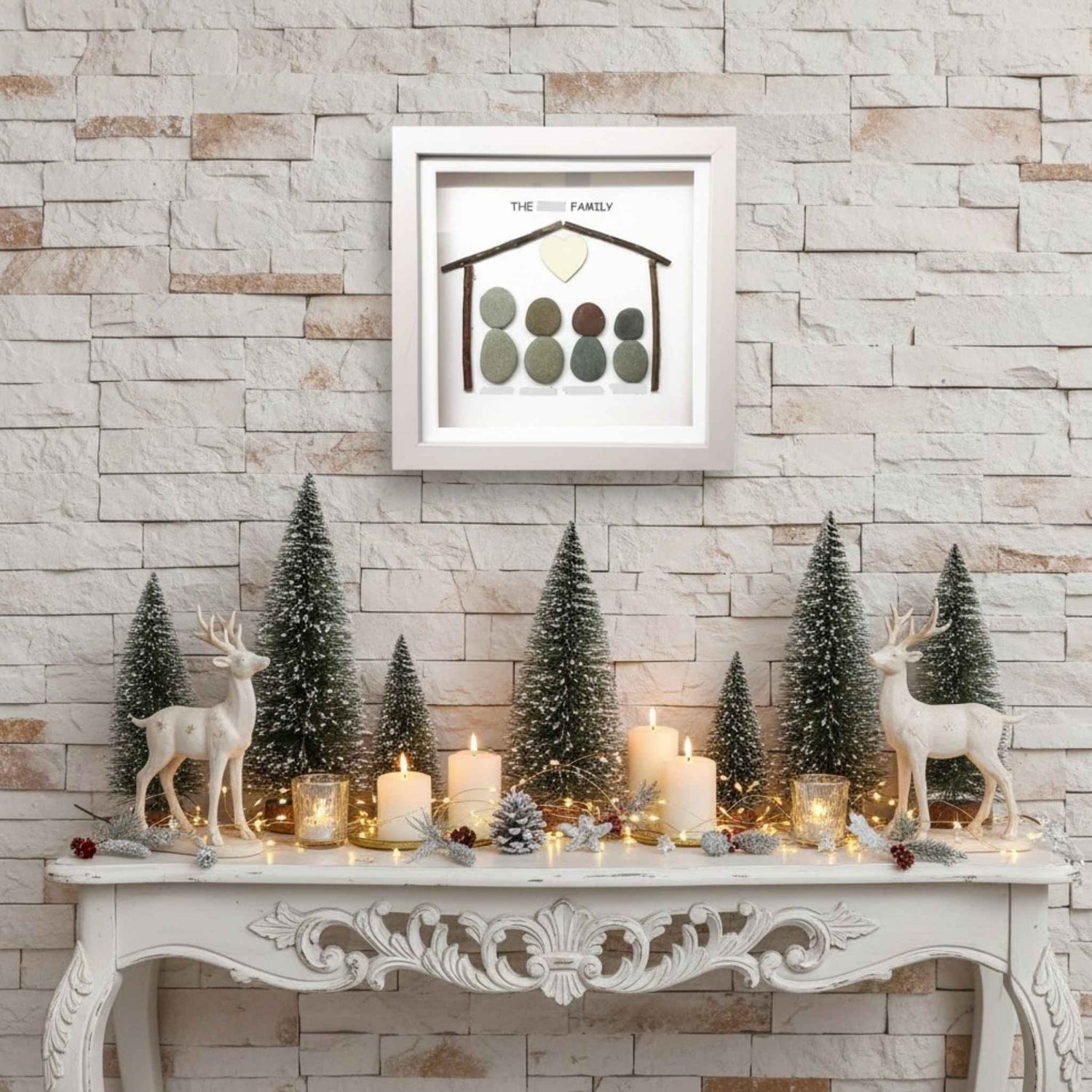 Decorative setup with Christmas trees, candles, and deer figurines on a mantelpiece against a stone wall. A pretty family pebble art frame is above.