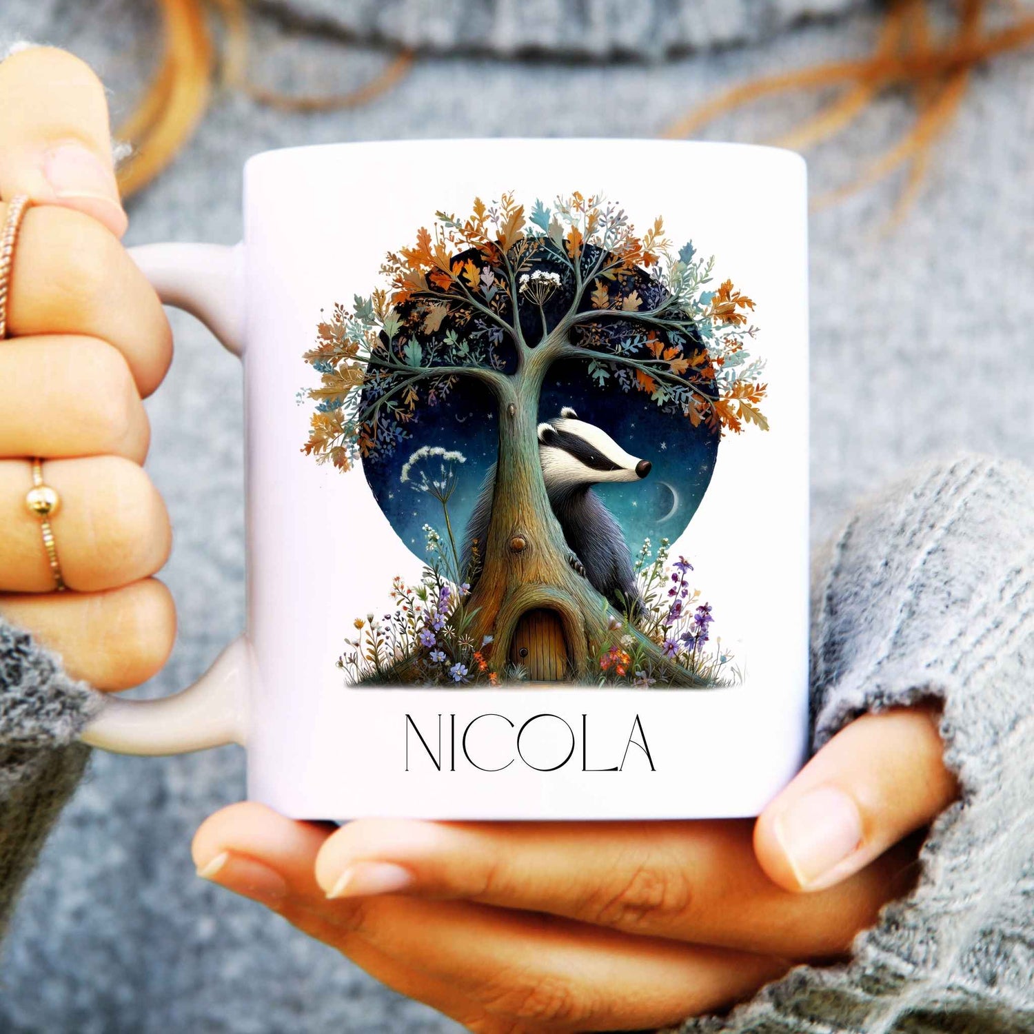 Person holding a mug with a tree and badger design, featuring the name 'Nicola'.