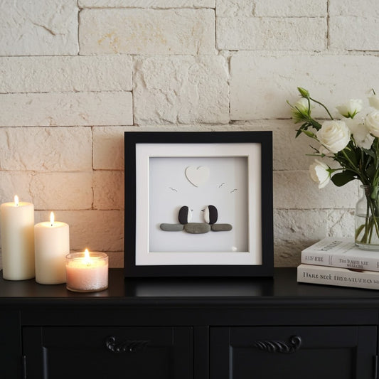 A framed minimalist art print featuring penguins, displayed on a black cabinet against a white brick wall, styled with candles, flowers, and books.
