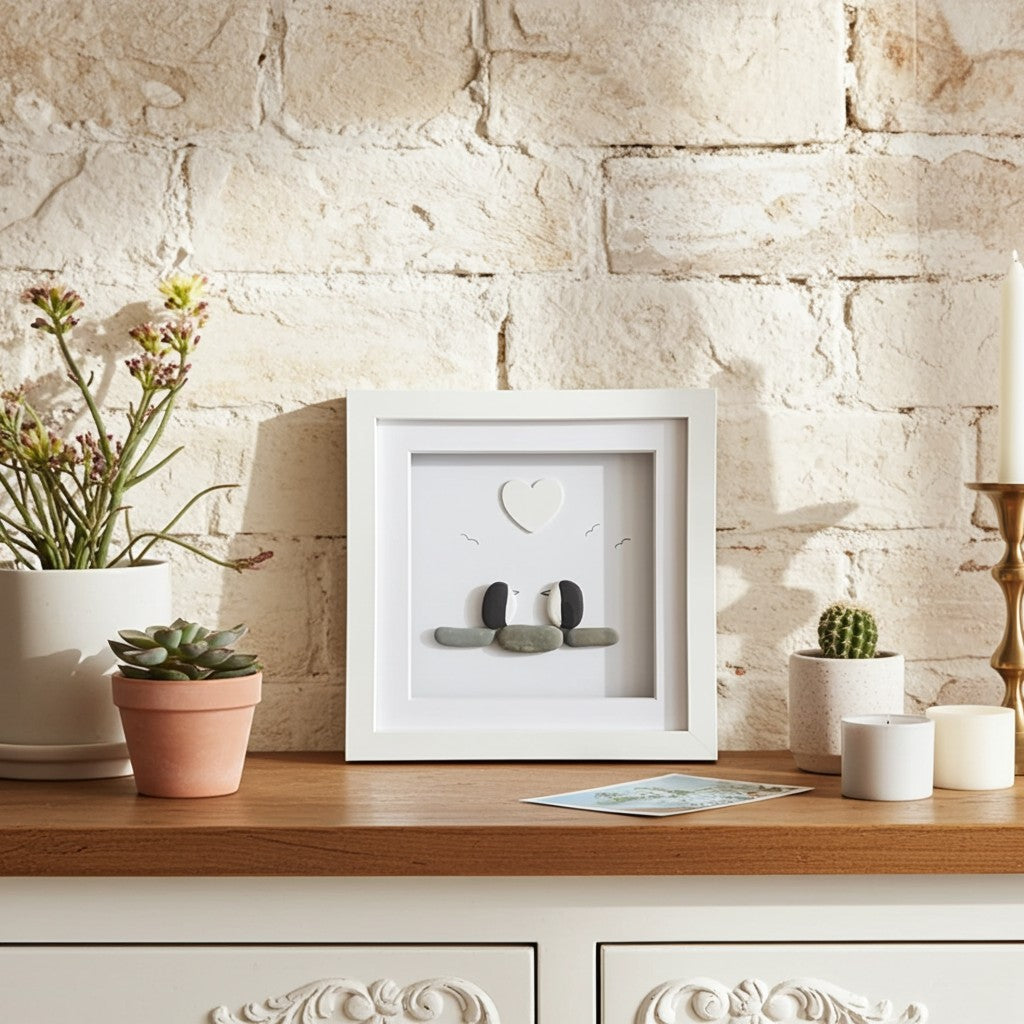 A white square shadow box frame with two penguins made from small stones and a heart detail, displayed on a wooden cabinet against a rustic white brick wall with potted plants and candles.