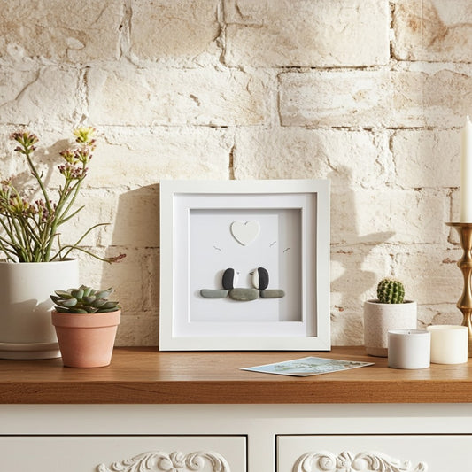 A white square shadow box frame with two penguins made from small stones and a heart detail, displayed on a wooden cabinet against a rustic white brick wall with potted plants and candles.